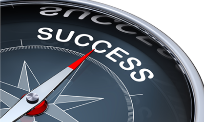 A dark gray compass with a chrome rim and a silver dial with a red tip pointed towards the word success