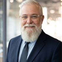Caucasian man with glasses, gray hair and a gray beard wearing a navy blazer with a light blue shirt and navy tie.