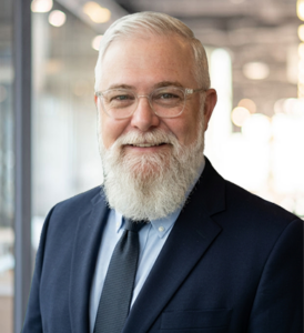 Caucasian man with glasses, gray hair and a gray beard wearing a navy blazer with a light blue shirt and navy tie.