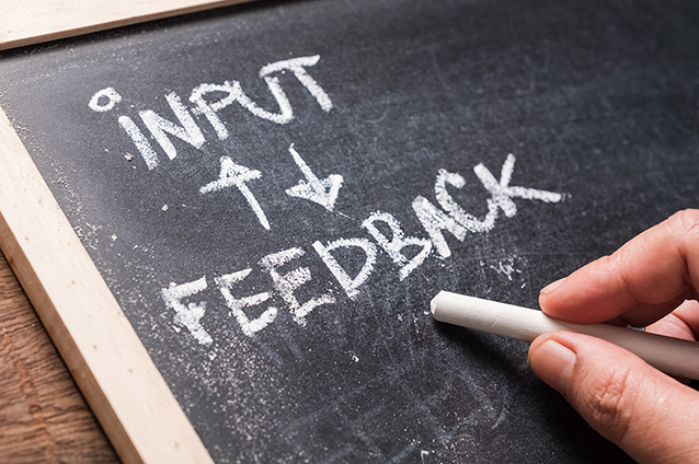Dark chalkboard with a light wood border with the words input and feedback written in white chalk with directional arrows between the words.